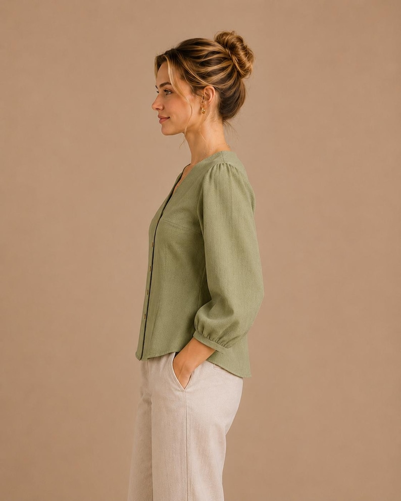 May include: A sage green long-sleeve button-down shirt with a V-neckline and gathered cuffs, paired with light beige trousers. The shirt features a straight hemline and a simple, clean design, showcasing a soft, textured fabric appearance.