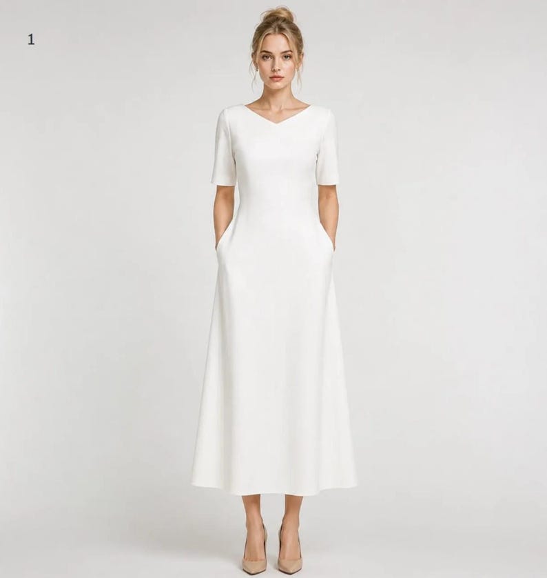 May include: A solid white midi-length dress featuring a V-neckline, short sleeves, and a fitted bodice that transitions into a flared A-line skirt. The garment includes two side pockets integrated into the seams of the skirt, providing a clean look.