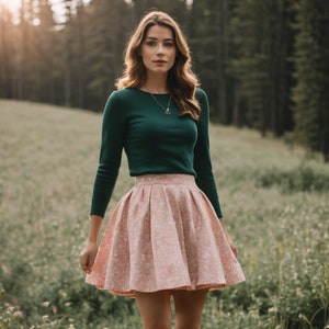 Könnte beinhalten: Eine Frau trägt einen dunkelgrünen Langarm-Pullover und einen rosa Plisseerock mit weißem Blumenmuster. Sie steht auf einer Wiese mit grünem Gras und Wildblumen.