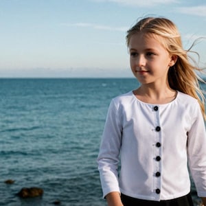 May include: A white long-sleeved cardigan with black buttons. The cardigan is worn by a person standing near the ocean. The person has blonde hair and is looking towards the right. The background includes the ocean and a shoreline.