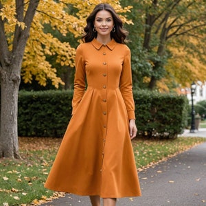 May include: A long-sleeved, button-down dress in a warm orange color. The dress has a collared neckline and a fitted bodice with a flared skirt. The dress is made of a smooth, woven fabric.