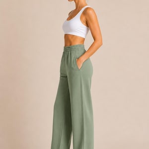 May include: A white racerback sports bra and sage green high-waisted sweatpants with a wide-leg silhouette, an elastic waistband, and side pockets. The sweatpants feature a vertical seam detail down the front of each leg and are paired with white sneakers.