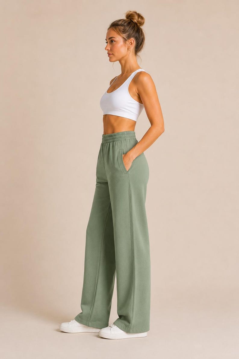 May include: A white racerback sports bra and sage green high-waisted sweatpants with a wide-leg silhouette, an elastic waistband, and side pockets. The sweatpants feature a vertical seam detail down the front of each leg and are paired with white sneakers.