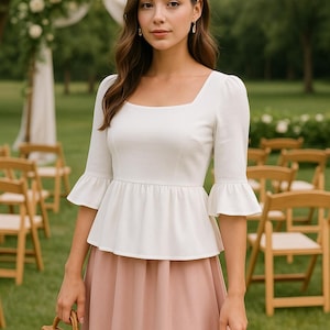 May include: A woman wearing a white, square-neck blouse with a ruffled peplum and bell sleeves. She is also wearing a blush-colored skirt and a woven wicker handbag.