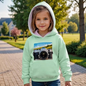 May include: A light green hoodie with a pink-lined hood. The front of the hoodie features a rectangular graphic of a vintage camera against a backdrop of a blue sky and green field. The girl is wearing the hoodie outside.