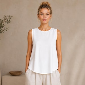 May include: A sleeveless white top with a round neckline and a relaxed, slightly flared fit. The garment features a clean, solid white color and a simple, unadorned design with a hemline that curves slightly at the sides for a soft silhouette.
