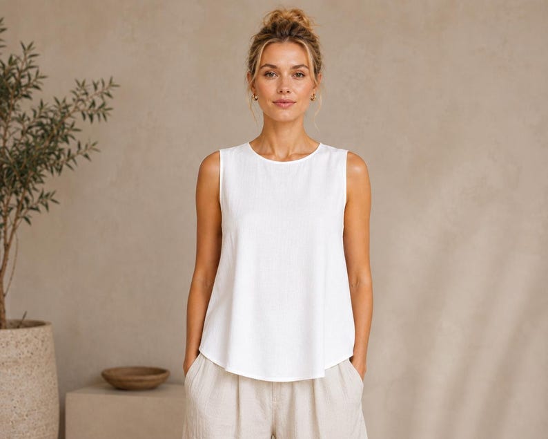 May include: A sleeveless white top with a round neckline and a relaxed, slightly flared fit. The garment features a clean, solid white color and a simple, unadorned design with a hemline that curves slightly at the sides for a soft silhouette.