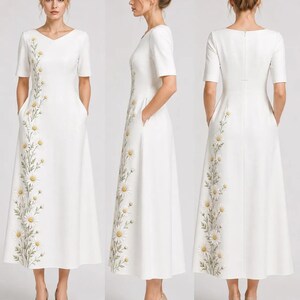 May include: A white midi dress featuring short sleeves, a V-neckline, and a vertical floral embroidery pattern of daisies with yellow centers and green stems running down the side. The dress includes side pockets and a concealed back zipper closure.