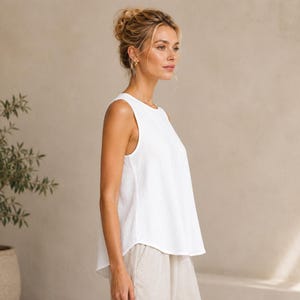 May include: A sleeveless white top with a round neckline and a relaxed, flared fit. The garment features a solid white color and a simple, clean design with a slightly curved hemline that falls at the hip, crafted from a lightweight fabric.