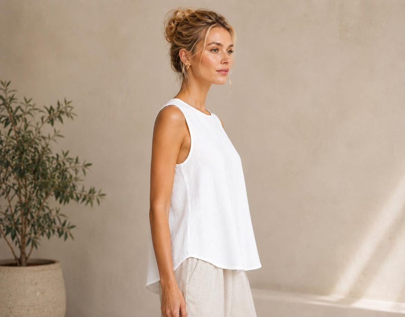 May include: A sleeveless white top with a round neckline and a relaxed, flared fit. The garment features a solid white color and a simple, clean design with a slightly curved hemline that falls at the hip, crafted from a lightweight fabric.