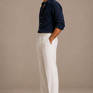 K&ouml;nnte beinhalten: Ein lang&auml;rmeliges, dunkelblaues Hemd mit Knopfleiste und Kragen, kombiniert mit einer wei&szlig;en Hose mit Faltenfront und Seitentaschen. Das Outfit wird durch braune Wildleder-Slipper und dunkel get&ouml;nte Sonnenbrillen erg&auml;nzt, getragen von einer Person.