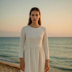 May include: An ivory-coloured dress with long sleeves, a round neckline, and a gathered waist. The dress falls to the knees. The person wearing the dress is standing on a beach, with the sea and sky visible.