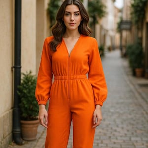 以下が含まれることがあります： 女性が、フロントにジッパーが付いた明るいオレンジ色のジャンプスーツを着ています。ジャンプスーツは長袖で、ウエストがシェイプされ、Vネックになっています。
