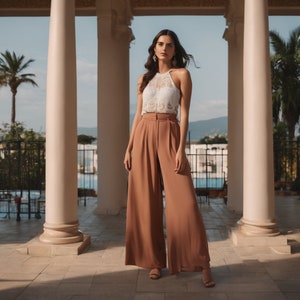 May include: A woman wearing a white lace halter top and wide-legged, rust-colored pants. She is standing in a white columned building with a view of a pool and palm trees in the background.