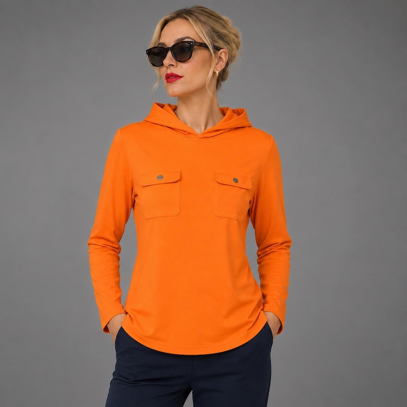 May include: A bright orange long-sleeve hooded pullover featuring two chest pockets with metallic buttons. The garment has a hood, a straight hemline, and long sleeves. The pullover is paired with dark navy blue trousers and black sunglasses.