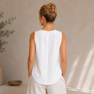 May include: A sleeveless white top featuring a round neckline and a vertical center seam on the back. The garment has a curved hemline and a concealed zipper closure positioned along the upper back, providing a clean and structured appearance.
