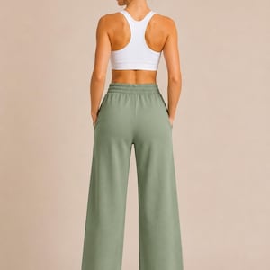 May include: A white racerback sports bra paired with sage green wide-leg sweatpants. The sweatpants feature a high-rise elastic waistband with a drawstring-free design and two side pockets. The fabric has a smooth, solid-colored finish.