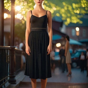 May include: A woman wearing a black midi dress with a thin gold belt. She is standing on a wooden platform with a railing behind her.