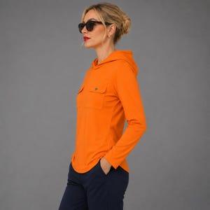 May include: A bright orange long-sleeve hooded shirt featuring two chest pockets with button closures, paired with dark navy blue trousers. The shirt has a hood and a straight hemline, worn with dark-rimmed sunglasses.