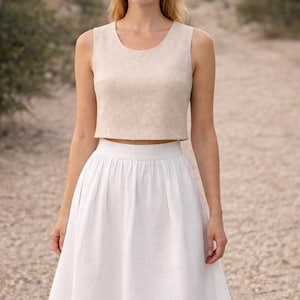 Op de afbeelding: Een beige mouwloze crop top en een witte midi rok. De top heeft een ronde halslijn en een getailleerd silhouet. De rok is hoog in de taille en uitlopend, gemaakt van een lichte stof. De outfit is geschikt voor warm weer en informele gelegenheden.