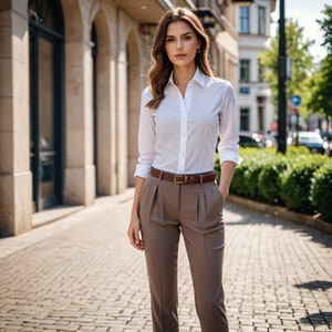 Op de afbeelding: Een vrouw draagt een wit overhemd met knopen, een bruine riem en een bruine broek. Ze staat voor een gebouw.
