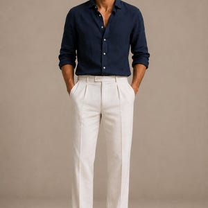 K&ouml;nnte beinhalten: Ein lang&auml;rmeliges, dunkelblaues Hemd mit Knopfleiste und Kragen, kombiniert mit einer cremewei&szlig;en, gefalteten Hose mit geradem Schnitt. Das Outfit wird durch braune Wildleder-Loafer und dunkel get&ouml;nte Sonnenbrillen abgerundet.