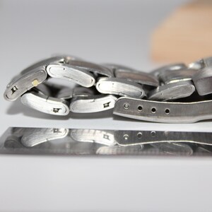 May include: Close-up of a silver-toned wristwatch with a metal band. The watch band is partially unclasped, revealing the intricate links and clasp mechanism. The watch face is visible, with a light-colored dial.