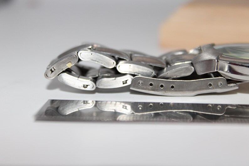 May include: Close-up of a silver-toned wristwatch with a metal band. The watch band is partially unclasped, revealing the intricate links and clasp mechanism. The watch face is visible, with a light-colored dial.