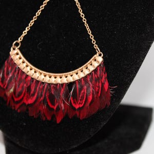 May include: A gold-toned necklace with a pendant featuring a fan of red and black feathers. The pendant has a curved, decorative gold metal top. The necklace is displayed on a black surface.