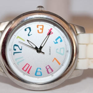 May include: A silver-toned quartz watch with a white face and a white, segmented band. The watch face features colorful numerals and black hands. The watch is a stylish accessory.