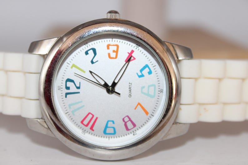 May include: A silver-toned quartz watch with a white face and a white, segmented band. The watch face features colorful numerals and black hands. The watch is a stylish accessory.