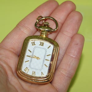 May include: A gold-toned rectangular pocket watch with a white face and black Roman numerals. The watch has a small loop at the top for attaching to a chain. The hands are gold, and the watch is held in a hand.