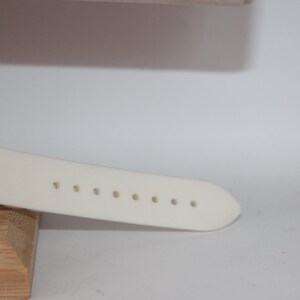 May include: Close-up of a cream-colored watch strap with evenly spaced holes. The strap is attached to a gold-toned watch face. The watch is resting on a wooden block, with a white background.