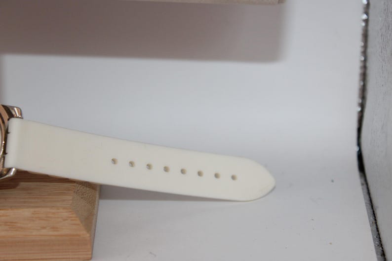 May include: Close-up of a cream-colored watch strap with evenly spaced holes. The strap is attached to a gold-toned watch face. The watch is resting on a wooden block, with a white background.