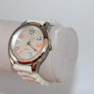 May include: A white wristwatch with a silver-colored bezel and a light-colored face. The watch face features colorful numbers and hands. The watch is on a white band and is displayed on a white cylindrical object.
