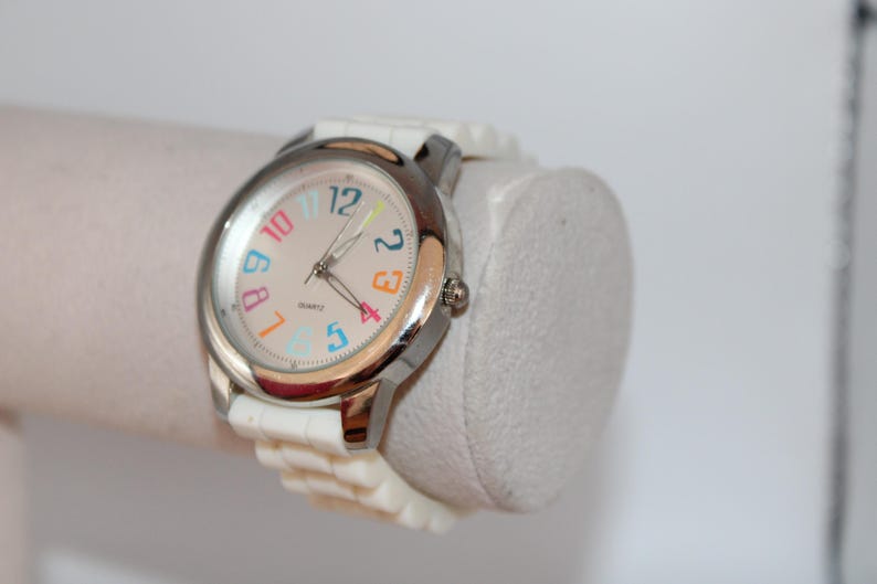May include: A white wristwatch with a silver-colored bezel and a light-colored face. The watch face features colorful numbers and hands. The watch is on a white band and is displayed on a white cylindrical object.