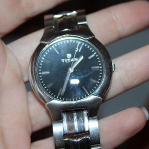 May include: A silver-toned Titan wristwatch with a black dial. The watch features a round face with silver hands and markers. The watch is held in a hand, showcasing the metal band and the watch's overall design.