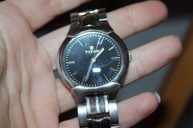 May include: A silver-toned Titan wristwatch with a black dial. The watch features a round face with silver hands and markers. The watch is held in a hand, showcasing the metal band and the watch's overall design.
