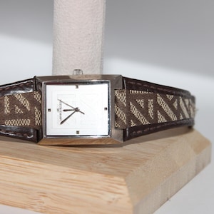 May include: A silver-toned Anne Klein watch with a square face and a brown leather strap. The watch face is white with black hands and markers. The strap features a brown and tan geometric pattern.