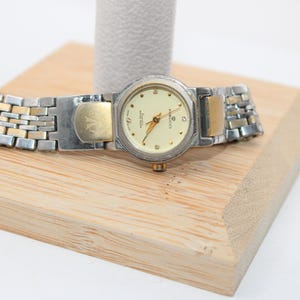 May include: A silver and gold-tone wristwatch with a cream-colored face. The watch has a rectangular face with gold hands and small diamond accents. The watch band is a combination of silver and gold links. The watch is displayed on a wooden stand.