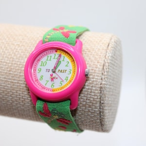 May include: A pink Timex watch with a green floral patterned strap. The watch face is white with pink and yellow accents, and the numbers are green. The strap is green with red and pink floral designs.