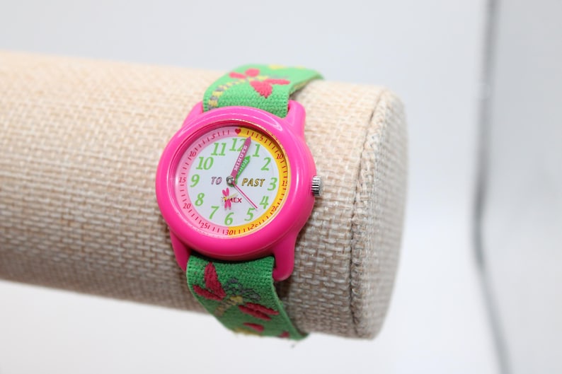 May include: A pink Timex watch with a green floral patterned strap. The watch face is white with pink and yellow accents, and the numbers are green. The strap is green with red and pink floral designs.