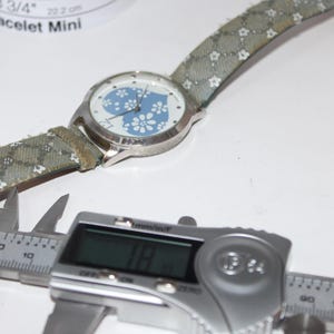 May include: A silver wristwatch with a blue floral design on the face and a patterned strap. The watch is next to a digital caliper displaying a measurement of 18 mm. The watch strap is approximately 8 3/4 inches or 22.2 cm.