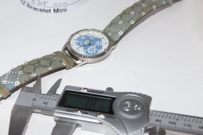 May include: A silver wristwatch with a blue floral design on the face and a patterned strap. The watch is next to a digital caliper displaying a measurement of 18 mm. The watch strap is approximately 8 3/4 inches or 22.2 cm.