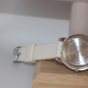 May include: A silver-toned wristwatch with a white band and a silver buckle. The watch is resting on a wooden surface. The back of the watch has text that is partially visible.