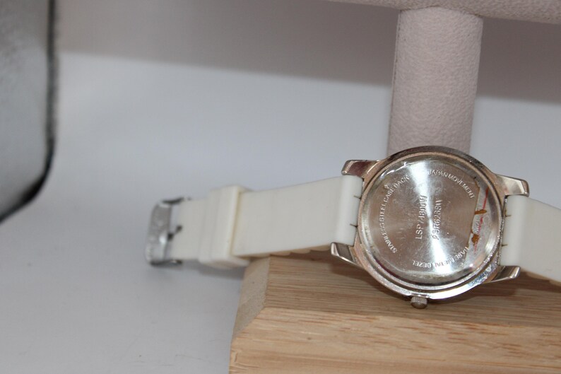 May include: A silver-toned wristwatch with a white band and a silver buckle. The watch is resting on a wooden surface. The back of the watch has text that is partially visible.