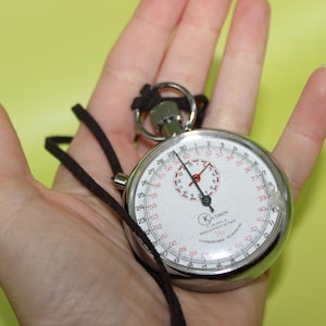 May include: A silver stopwatch with a white face, black numbers, and red accents. The stopwatch has a black cord and a metal ring. The watch face displays the brand name "Kalthron".