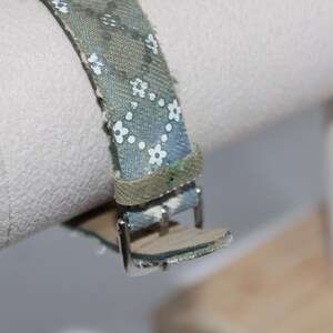 May include: Close-up of a watch strap with a blue and green floral pattern. The strap is made of fabric and has a silver buckle. The watch strap is attached to a white cylindrical object.
