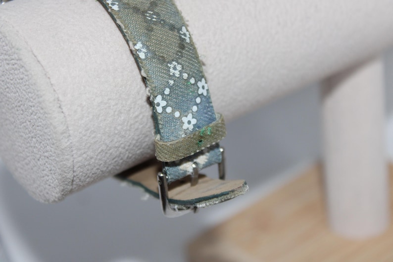 May include: Close-up of a watch strap with a blue and green floral pattern. The strap is made of fabric and has a silver buckle. The watch strap is attached to a white cylindrical object.