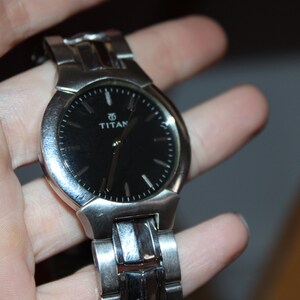 May include: A silver-toned Titan wristwatch with a black watch face. The watch has a round face with silver hour markers and hands. The watch band is also silver and appears to be made of metal.